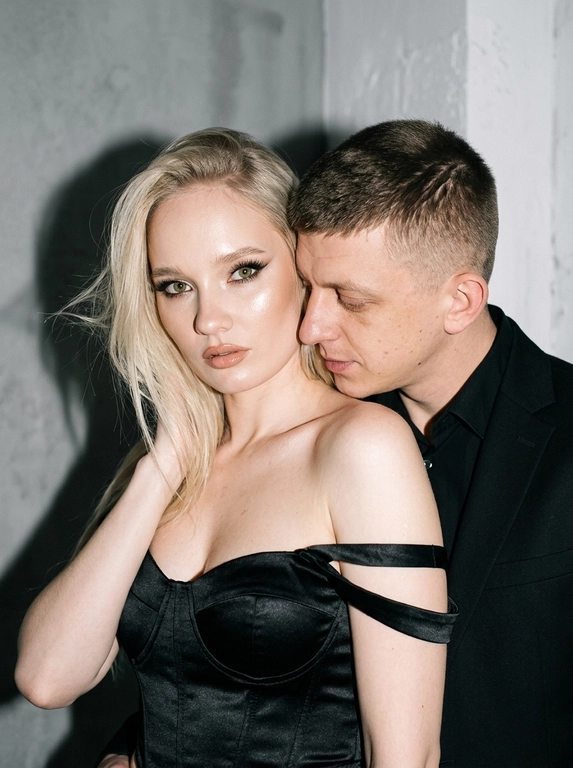 Не меняя черты лица, студийная fashion-съемка пары в минималистичном интерьере с серо-белым фоном, атмосфера гламура и интимной близости, ракурс — крупный портрет по грудь с легким поворотом корпуса, женщина на переднем плане, мужчина позади склоняется к её плечу, поза — она слегка отвернута от камеры с прямым взглядом, он касается лицом её шеи/плеча, одежда — на женщине черный корсет/бандо с открытыми плечами, на мужчине классический черный пиджак и рубашка, макияж — glam (ровный сияющий тон, сильный хайлайтер, четкий контуринг, стрелки, густые ресницы, глянцевые губы), свет — жесткая фронтальная вспышка + мягкий заполняющий свет, выраженные тени позади, эффект flash photography / editorial, холодно-нейтральная цветокоррекция с контрастом и глянцем кожи, высокая детализация, slight film grain, shallow depth of field, съемка на Canon EOS R / 50mm, f/2.0, direct flash, studio look.