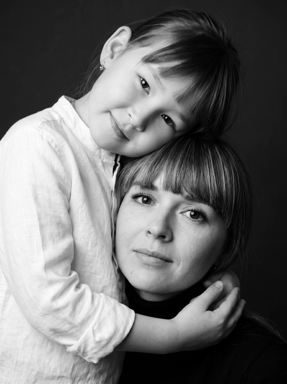 Родитель и ребёнок с внешностью как на приложенных фото, художественная семейная студийная фотосессия. Композиция — вертикальный портрет, крупный план лиц. Родитель внизу смотрит прямо в камеру, лицо фронтально. Ребёнок сверху, голова повернута набок, щека лежит на голове родителя, смотрит в камеру с нежной улыбкой. Рука ребёнка обнимает родителя за шею, видна в кадре, родитель держит руку ребёнка своей рукой.
Одежда: ребёнок в белой льняной рубашке, родитель в чёрной водолазке. 
Фон — тёмно-серый почти чёрный однотонный студийный, создаёт контраст. Освещение — мягкое боковое студийное, создаёт объём и глубину.
Чёрно-белая художественная фотография с выразительными контрастами. Атмосфера нежная, интимная, спокойная.