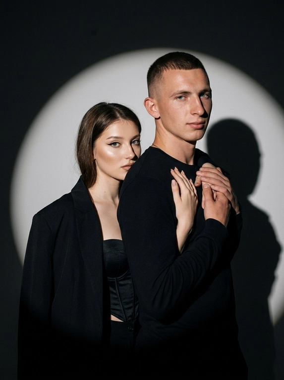 Фотореалистичный студийный портрет пары в драматичной Y2K fashion-эстетике. Мужчина стоит на переднем плане, ближе к камере, снят по грудь, крупнее в кадре. Его корпус слегка повернут в пол-оборота, одно плечо ближе к объективу, поза спокойная и уверенная. Женщина стоит позади него, немного ниже по уровню за счёт наклона корпуса, частично скрыта его плечом. Она мягко обнимает его за шею и плечо, её руки лежат спереди у его груди и ключиц. Мужчина одной рукой аккуратно держит её кисть. Её лицо видно из-за его плеча, близко к нему, поза выглядит интимно, дерзко и editorial, без чрезмерной романтичности. Женщина выглядит выразительно и гламурно: cat-eye стрелки, губы с чётким контуром, хайлайтер на коже, чёрный корсет и оверсайз-пиджак с открытым плечом. Мужчина одет в чёрный лонгслив. Фон студийный, тёмно-серый, почти чёрный. За парой на стену направлен мощный прожектор, создающий большой круг света позади их силуэтов. Это именно свет от прожектора, падающий на фон, а не нарисованное пятно: круг света мягко размывается по краям и постепенно уходит в темноту. Персонажи частично перекрывают свет от прожектора, поэтому на фоне видны реалистичные жёсткие тени от их тел, рук и плеч. Края кадра затемнены, вокруг светового круга глубокая тёмная виньетка, создающая ощущение сценического света. Освещение на персонажах: жёсткая фронтальная вспышка в Y2K-стиле, высокий контраст, реалистичные блики на коже, глубокие тени, лёгкое плёночное зерно. Камера на уровне глаз, портретный объектив около 100 мм, плотное кадрирование, 8K RAW, натуральная текстура кожи, видимые поры, реалистичные тени и блики, editorial fashion photo, без текста, без логотипов, без лишних предметов в кадре.