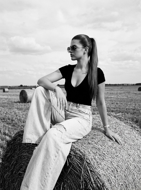 Чёрно-белая high-fashion editorial фотография в сельской местности, женщина с внешностью как на референсе, черты лица не изменять, композиция — более общий план (wide shot), камера на среднем расстоянии, фигура полностью в кадре с окружающим пространством, она сидит на большом тюке сена в поле, корпус слегка развернут, одна нога свободно опущена вниз, вторая согнута, создавая расслабленную, но выразительную позу, руки естественно лежат на колене и на поверхности тюка, взгляд направлен в сторону, профиль, выражение спокойное и уверенное, одежда: чёрный топ с V-образным вырезом и объёмные светлые широкие джинсы с потёртостями и рваными элементами, аксессуары: крупные солнцезащитные очки, минималистичные украшения, фон: открытое поле с несколькими тюками сена на разной глубине, создающими перспективу, линия горизонта ниже центра кадра, чтобы подчеркнуть небо, освещение: жёсткий естественный дневной свет с мягкими облаками, формирует выразительные тени и контраст, стиль: кинематографический, плёночное зерно, высокая детализация, реалистичная текстура кожи и ткани, редакционный fashion стиль, акцент на атмосфере и пространстве кадра


Какая будет прическа?: Гладкие волосы собраны в высокий хвост