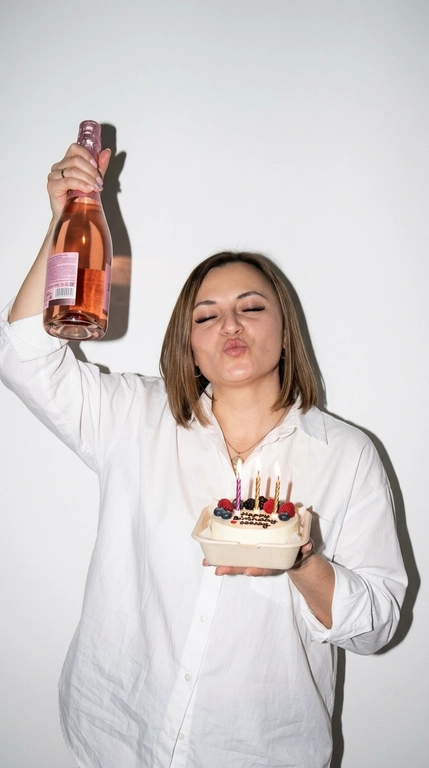Ультрареалистичная фотография в эстетике Party Girl / Birthday Aesthetic, стилизация под случайный снимок на компактную цифровую камеру начала 2000-х.
 девушка празднует день рождения. На ней свободная белая рубашка оверсайз на пуговицах. В одной руке она высоко держит бутылку розового шампанского, в другой — небольшой бенто-торт со светлыми свечами и свежими ягодами.

Глаза закрыты, на лице игривое выражение с лёгким «поцелуйным» жестом губ. Не меняй прическу модели.

Съёмка со вспышкой прямо в камеру на фоне простой молочно-белой стены. Жёсткий свет создаёт характерную чёткую тень за фигурой, яркие блики на коже, волосах, бутылке и глазури торта. Атмосфера спонтанного домашнего праздничного снимка: немного несовершенная композиция, лёгкая зернистость, естественная текстура кожи, реалистичные отражения и мелкие детали.

Стиль: 2000s digital camera, lo-fi party aesthetic, candid snapshot, hard flash, authentic lifestyle photography, natural skin texture, realistic colors, high detail, 4K.

Без: студийной постановки, мягкого идеального света, пластиковой кожи, чрезмерной ретуши, CGI, мультяшности, искажённых рук и пальцев, лишних пальцев, деформированной бутылки или торта, артефактов текста, логотипов и водяных знаков.

Доп. характеристики: Сделать лицо похудее и нарастить ресницы девушке