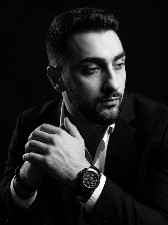 A hyper-realistic 8K cinematic portrait of a uploaded person,
captured in a dramatic Chiaroscuro style. The subject is seated at a
three-quarter angle, leaning slightly forward with a relaxed yet
commanding posture. His face is turned away from the lens, with
one side of his face clearly illuminated while the other is
completely lost in deep, velvety black shadows. His expression is
contemplative, not looking at the camera. The pose is precise: his
hands are clasped together near his chest; one hand is held
straight with fingers slightly tilted, while the other hand is
positioned so only the fingers are visible, resting over the first. He
wears a luxury black chronograph watch with a detailed metal link
bracelet, and a beaded bracelet. His attire is a sharp
black suit jacket over a white dress shirt with the top buttons
open, showing refined fabric textures. The background is a solid,
seamless black. The lighting is sharp and directional, highlighting
the intricate details of his hair,
the moisture in his eyes, and the glint off the watch face.
Cinematic black-and-white color grade with rich contrast and
smooth, natural skin transitions. Shot on 85mm lens, f/1.8, high-
resolution commercial photography
