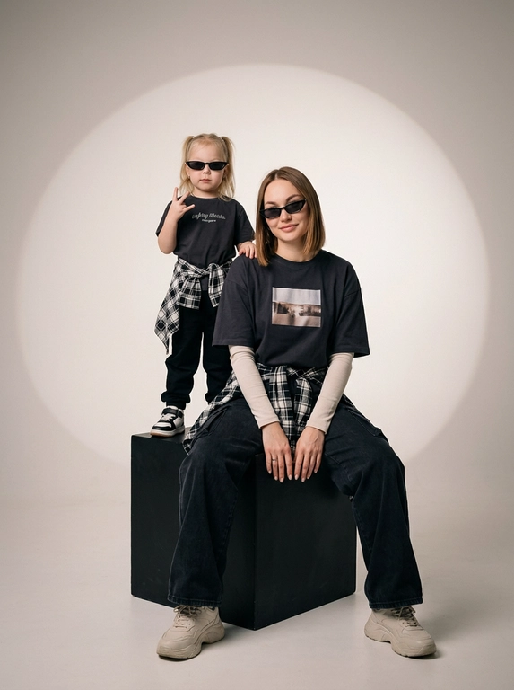 Photorealistic studio portrait photography in minimalistic fashion/editorial aesthetics, static camera at chest level of a seated person, light frontal angle without noticeable perspective deformation; composition with two characters: an model sits on a large matte black cube, body slightly turned towards the camera, knees bent, feet on the floor, the arms are lowered and relaxed in front of the knees, the fingers are neatly extended; the child stands on the same cube behind and to the left, at full height, one hand raised with a "V" gesture, the other lowered, pose confident and playful; both wearing narrow dark sunglasses, gaze directed at the camera; shooting plan — full height for both with a small supply of air above and below, center the center of the frame is shifted to the right to a sitting figure, a standing child balances on the left; the lens is normal/short portrait 50-70 mm, without width; aperture is approximately f/5.6–f/8, moderate depth of field, both characters are sharp, the background is slightly soft; the studio lighting is soft, with one key source on the front left at a level just above the head (softbox), giving smooth transitions and a pronounced but soft shadow against the background on the right; light filling light from the front reduces contrast, shadows are readable; the background is a smooth, light, seamless cyclorama/wall with a noticeable large oval light spot (gradient) in the center, there is a darker vignetting around, a clean minimalistic studio without props; the mood is calm, stylish, slightly retro, with an emphasis on street fashion and family vibe; the expression on the face of the seated model is a soft smile, relaxation; the person standing has a confident neutral facial expression; clothes: the seated model has a dark oversized T-shirt with a large rectangular print on the chest, under it a light longsleeve with long sleeves, wide dark cargo trousers/joggers with pleats at the knees and bottom, a plaid shirt/flannel tied at the waist with a knot, massive light sneakers with a pronounced sole and mesh texture; the child has a dark T-shirt with a large inscription on the chest, dark trousers, a plaid shirt tied at the waist, sneakers in two colors; fabrics with a realistic cotton and flannel texture, noticeable fine folds, natural creases on the knees and in the waist area, matte surfaces without gloss; micro-details: natural skin texture without plastic, neat glare on the lenses of glasses, a clear contour of the shadow from the raised hand on the background, clean floor and soft drop shadow from the cube; high detail, natural sharpness, light grain like a film shot, moderate contrast, soft matte/film look, no HDR, no overexposure and no unnecessary objects.

Какого цвета будут рубашки на поясе?: Чёрно-белый.