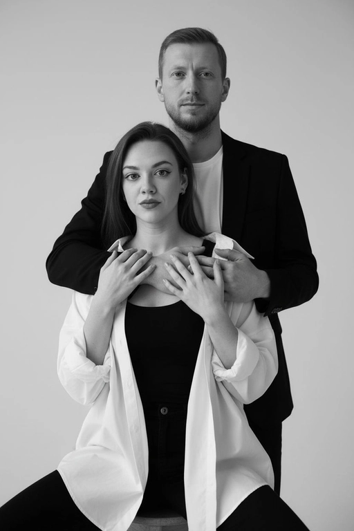 Черно-белый портрет мужчины и женщины в стиле фэшн фотосессии в фотостудии. Не изменяйте лица. Фон нейтральный, однотонный, светло-серый. Атмосфера интимная и спокойная. На переднем плане женщина. Вся композиция выполнена в приглушенной монохромной гамме со студийным освещением, которое подчеркивает текстуру кожи и одежды. Её руки лежат на груди мужчины. Мужчина обнимает женщину сзади, положив свою руку поверх её руки. Она одета в черный топ, белую оверсайз рубашку. Мужчина одет в  классическом черном пиджаке и белой футболке.