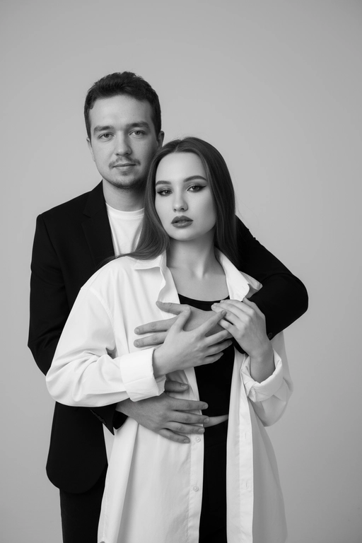Черно-белый портрет мужчины и женщины в стиле фэшн фотосессии в фотостудии. Не изменяйте лица. Фон нейтральный, однотонный, светло-серый. Атмосфера интимная и спокойная. На переднем плане женщина. Вся композиция выполнена в приглушенной монохромной гамме со студийным освещением, которое подчеркивает текстуру кожи и одежды. Её руки лежат на груди мужчины. Мужчина обнимает женщину сзади, положив свою руку поверх её руки. Она одета в черный топ, белую оверсайз рубашку. Мужчина одет в  классическом черном пиджаке и белой футболке.
