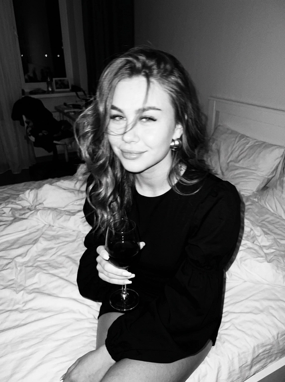 Create a portrait, transfer the face from the reference photo without changing facial features, proportions, or expression, preserve full identity. Aesthetic black-and-white nighttime Y2K photograph captured with direct on-camera flash, shot in motion.
A woman is sitting on the edge of a white, slightly rumpled bed, the frame taken from a slightly elevated angle. She wears a stylish black top with voluminous sleeves and large round statement earrings. In one hand she holds an elegant glass of dark wine. Her body is subtly moving, creating natural motion blur across the face and shoulders, as if the moment was caught mid-gesture. The gaze is languid and directed toward the camera, with a soft, intimate smile. Hair falls in large waves with loose strands drifting across the face, slightly displaced by movement. Lighting is harsh flash against deep night shadows, high contrast with blown highlights and strong falloff, visible film grain. Skin texture remains real and alive, no smoothing, enhanced by flash glare. The mood is high-fashion editorial, Parisian chic, old-money sensuality, nighttime melancholy, spontaneous and cinematic, shot in a raw 35mm film aesthetic with pronounced motion blur and lived-in realism.