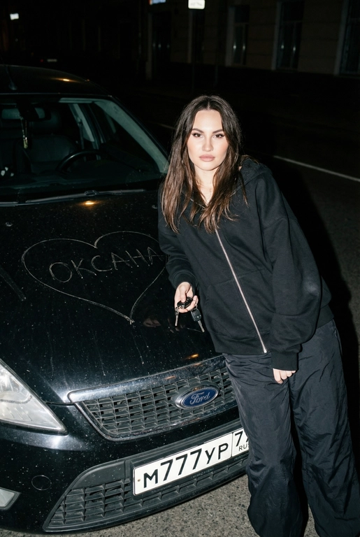 Строго сохранить лицо с прикреплённой референс-фотографии, на 100% без искажений. Человек одет полностью в чёрный, монохромный оверсайз спортивный костюм: свободная худи на молнии и широкие расслабленные штаны, без логотипов, текста и брендинга. Акцент на реальной текстуре кожи и детализированной текстуре ткани.
Сырой кинематографичный ночной кадр. Человек опирается на капот автомобиля, средний план, лёгкий верхний ракурс. В одной руке игриво держит ключи от машины. Номерной знак виден: m777yp 77.
Строго по центру капота, покрывая всю поверхность капота, глубоко и грубо процарапаны (не мелом) царапины: большое сердце с текстом «» внутри.
Жёсткая прямая вспышка, сильные тени, высокий контраст, видимая пыль и микро-царапины на краске. Тёмный размытый городской фон.
Объектив 35mm, f/2.8, грубая уличная эстетика.

Надпись на капоте: Оксана

Марка автомобиля: Форд мондео