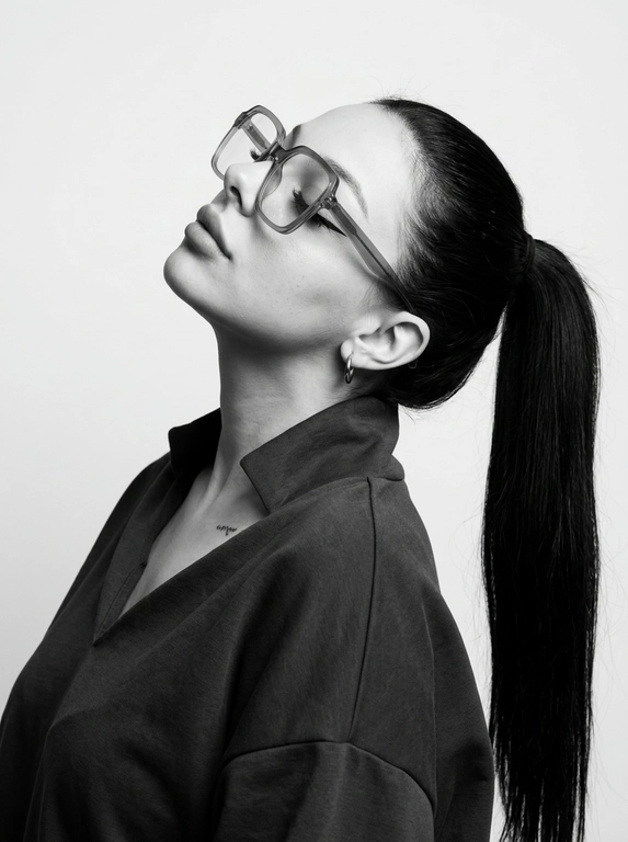 Чёрно-белый минималистичный фотореалистичный портрет, студийный мягкий свет спереди‑сверху, нейтральная светлая однотонная стена как фон. Девушка в профиль: голова запрокинута назад, подбородок поднят, глаза закрыты — расслабленная fashion-поза с акцентом на линии шеи и контуры лица. Обязательно точно сохранить черты лица с референс-фото (без искажений); крупные квадратные очки в массивной полу-прозрачной оправе с естественными бликами. Детали лица: длинные ресницы и чёткие стрелки, выразительные губы, естественная текстура кожи, аккуратный ретуш. Одежда: тёмный объёмный верх с выразительным воротником, маленькая серьга-кольцо. Стиль: высокая графичность и контраст, чистые линии, лёгкая драматичность, максимальная детализация и фотореализм, аккуратные светотеневые переходы, подчёркнутые контуры.

Какая будет прическа?: Гладкие волосы, высокий длинный густой хвост.