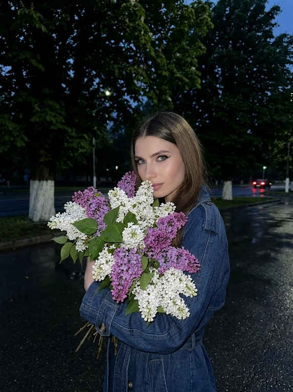 Очень крупный план, на этом фото женщина стоит в анфас к камере, обеими руками она прижимает к себе огромную пышную охапку сирени белой и фиолетовой, её голова слегка наклонена к цветам создавая ощущение что она вдыхает их аромат смотря в камеру, макияж: глаза подчеркнуты чёрными стрелками и длинными ресницами, на губах блеск, фон: действие происходит на улице в глубоких сумерках или ночью на заднем плане видны тёмные массивные силуэты деревьев с густой майской листвой внизу выкрашенные в белый цвет земля под ногами выглядит как тёмный асфальт после дождя на заднем плане едва заметные красные блики возможно огни далёких машин, ракурс: съёмка ведётся со стороны чуть сбоку на уровне глаз, освещение: слабое ночное освещение, фотореализм высокая детализация фото снято на камеру iPhone 17 Pro эффект живого кадра лёгкий цифровой шум зернистость мягкая резкость высокая детализация, без вспышки.

Какая будет одежда?: Темно-синий джинсовый тренч
