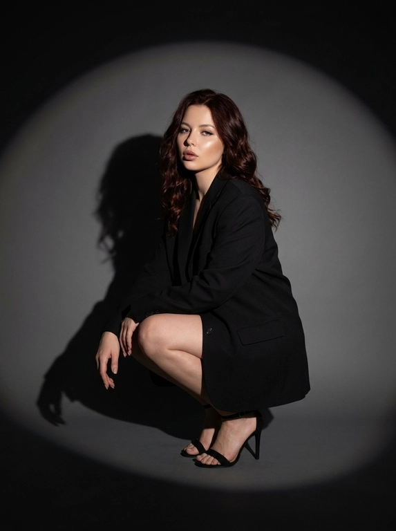 Модная студийная съёмка в стиле fashion editorial. Не менять внешность женщины. Женщина одета в только в чёрный оверсайз пиджак. Профессиональный макияж с акцентом на кожу и глаза. Один направленный прожектор формирует чёткое круглое световое пятно на темно-сером бесшовном гладком фоне и выразительную тень за спиной. Контрастный кинематографичный свет, глубокие тени, драматичный минимализм. Атмосфера — уверенность, сила, эстетика high fashion. высокая детализация кожи, реалистичная текстура, съёмка уровня модного журнала. Средний план. Женщина сидит на корточках, одна рука лежит на колене, корпус слегка наклонён вперёд. Уверенный прямой взгляд в камеру. На ногах чёрные босоножки на тонком каблуке. Чёткая тень позади усиливает драматизм кадра.

Какая будет прическа?: Волосы распущены, уложены в легкие волны.