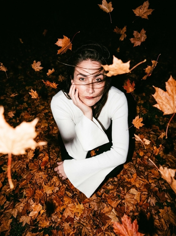 Ultra-realistic candid NIGHT autumn photo. The woman sits curled up directly in a thick carpet of dry autumn maple leaves, knees pulled toward her chest. Camera positioned very high above her, almost directly overhead, dramatic top-down perspective. She raises her face toward the camera, one hand gently supporting her cheek, the other arm resting across her knees.
She wears a fitted white knitted top with very long dramatic FLARED BELL SLEEVES, fitted through the arms and strongly widening from the elbows.
Strong autumn wind blows her hair.

It is DARK outside, almost nighttime. The background beyond the flash falls into deep natural darkness. Shot with a powerful direct on-camera flash: her face, white sweater and the closest leaves are brightly illuminated, while distant leaves rapidly disappear into black shadows. Dozens of dry orange, rust and brown leaves are FLYING THROUGH THE AIR around her, caught mid-flight by the flash; some pass very close to the lens and create strong realistic motion blur and streaks. A few strands of hair also have motion blur from the wind. Face remains sharp. Raw spontaneous flash photography, high contrast, deep blacks, warm saturated autumn tones, slight grain, imperfect handheld framing, realistic textures, no studio lighting, no HDR, no AI look.