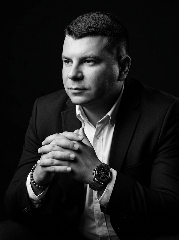 A hyper-realistic 8K cinematic portrait of a uploaded person,
captured in a dramatic Chiaroscuro style. The subject is seated at a
three-quarter angle, leaning slightly forward with a relaxed yet
commanding posture. His face is turned away from the lens, with
one side of his face clearly illuminated while the other is
completely lost in deep, velvety black shadows. His expression is
contemplative, not looking at the camera. The pose is precise: his
hands are clasped together near his chest; one hand is held
straight with fingers slightly tilted, while the other hand is
positioned so only the fingers are visible, resting over the first. He
wears a luxury black chronograph watch with a detailed metal link
bracelet, and a beaded bracelet. His attire is a sharp
black suit jacket over a white dress shirt with the top buttons
open, showing refined fabric textures. The background is a solid,
seamless black. The lighting is sharp and directional, highlighting
the intricate details of his hair,
the moisture in his eyes, and the glint off the watch face.
Cinematic black-and-white color grade with rich contrast and
smooth, natural skin transitions. Shot on 85mm lens, f/1.8, high-
resolution commercial photography