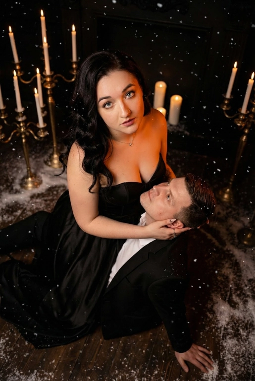 Ракурс камеры чуть сверху и сбоку. Романтическая и драматичная сцена в тёмном помещении. На полу сидит пара — мужчина и женщина верхом на мужчине в тесном, интимном положении. Женщина повернута к камере, её лицо поднято вверх, взгляд в камеру, хорошо освещено мягким тёплым светом. Она смотрит прямо вперёд, немного приоткрыв губы, с выразительным, глубоким взглядом. Если у женщины на фото длинные волосы. то они уложены в роскошные голивудские волны. Если короткие, то волосы просто красиво лежат.  На ней женщине длинное черное платье с лифом-бюстье без бретелей, подчеркивающее силуэт, создающими драматический силуэт.

Мужчина сидит у её груди, склонив голову к ее ключице и смотрит в ее лицо снизу вверх, словно обнимая её, одной рукой. Другой рукой упирается в пол позади себя. Он в чёрном классическом костюме и белой сорочке. Женщина одной рукой держит его за затылок, пальцы мягко погружены в волосы, создавая ощущение близости и страсти.

Вокруг пары падают искусственные снежинки — мелкие белые хлопья, подсвеченные тёплым светом свечей, что создаёт атмосферу зимней магии и романтики. На полу вокруг них разбросан «снег», добавляя глубины сцене.

Позади пары стоят золотистые и латунные канделябры с горящими свечами — тонкие высокие свечи создают мягкое золотое сияние. Из-за низкого освещения фон почти исчезает, оставляя только теплые огоньки и силуэты свечей. Освещение — контрастное: тёплый свет от свечей и мягкая направленная подсветка на лица. Очень темное фото.

Пол — тёмный, возможно деревянный, слегка блестящий от света. Атмосфера кадра — готическая, чувственная, тёплая, драматическая и немного мистическая.