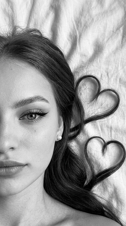 Фото снято на фронтальную камеру iPhone 16 Pro Max. Фотореалистичное чёрно-белое селфи девушки, лежащей на светлой слегка смятой простыне. крупный план. Камера расположена прямо над лицом. В кадре видна только половина лица, один глаз смотрит прямо в камеру.

Макияж выразительный и аккуратный, в эстетике clean soft glam: чёткие графичные чёрные стрелки с вытянутым эффектом, длинные густые ресницы с лисьим эффектом, хорошо оформленные брови с аккуратной укладкой наверх, мягкий контуринг, деликатный хайлайтер во внутренних уголках глаз, на скулах и спинке носа, губы с чётким контуром и мягким гладким финишем. Макияж выглядит безупречно, дорого и очень чисто.

Волосы естественно уложены на одну сторону вокруг головы, две крупные пряди случайно образуют два сердца, остальные волосы свободно и натурально лежат волнами. Мягкий естественный дневной свет из окна, лёгкие тени. Высокая детализация лица, чётко видны поры кожи, текстура губ, ресницы, брови и отдельные волоски. Волосы максимально детализированные, с естественным блеском и объёмом. Минималистичный кадр, светлая ткань с натуральными складками на фоне, без лишних предметов. Фото снято на фронтальную камеру iPhone 16 Pro Max.