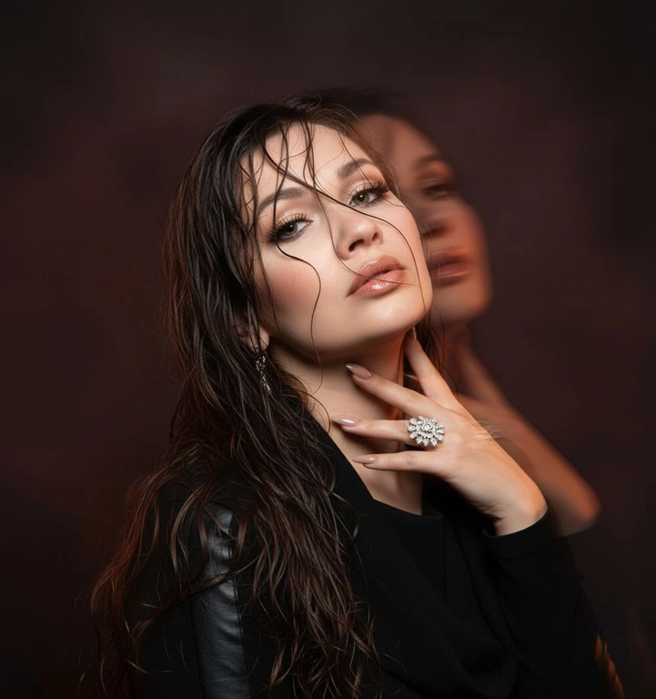 Создайте портрет женщины с длинными, волнистыми, влажными волосами, свободно ниспадающими на плечи, некоторые пряди прилипают к лицу. Она чувственно смотрит в камеру, слегка откинув голову назад, словно наслаждаясь мгновением покоя или чувственности. Её рука нежно лежит на шее, на пальце – эффектное сверкающее кольцо. Плечи опущены. Тёмный, почти однотонный и размытый фон подчёркивает силуэт и лицо женщины. Весь образ мастерски передан с помощью размытия в движении и световых бликов, которые создают ощущение естественного, сказочного движения головы и руки. Мягкие, тёплые тона, лёгкое свечение кожи и губ создают интимную, чувственную и таинственную атмосферу. Эффекты: размытие в движении, художественная длительная выдержка, тёплые тона, атмосфера сновидения. Макияж выразительный: длинные чёрные ресницы, бежевые тени с лёгким мерцанием, выразительные контуры и румяна, безупречная кожа и пухлые губы с нюдовым блеском и тёмно-коричневым карандашом для губ. Ухоженные брови с эффектом Bravenlifting. Длинные, заостренные ногти. Реалистичное фото, как будто снято на профессиональную камеру. Сохраните черты лица такими, какие они есть.