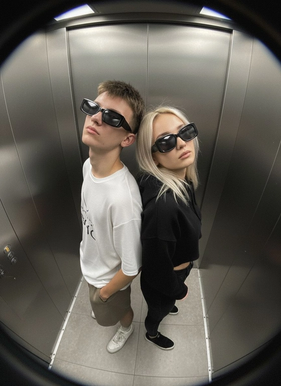Используй предоставленные изображения и создай фото с двумя людьми в лифте с металлическими стенами. Цветовая гамма с преообладанием серых металлических оттенков. Кадр снят с максимально верхнего ракурса, люди будто заглядывают в камеру, их головы подняты наверх, взгляд в камеру. Легкий эффект fish eye. Реалистичное фото. Зернистость пленочного фото, насыщенные цвета, стиль фото y2k. Сохрани черты лица людей. Они стоят спиной друг к другу. Руки в карманах. На них чёрные трендовые очки. Девушка в чёрных джинсах как на фото