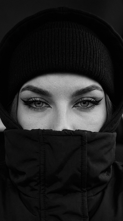 Не меняя внешности женщины с фото, перед нами крупный план её лица, частично скрытого под чёрной шапкой-бини, капюшоном и чёрной курткой с высоким воротом, которые обнимают нижнюю часть лица, оставляя видимыми только выразительные глаза.

Очень близкое селфи на iPhone 16, горизонтальный кадр, видно только глаза и брови, взгляд направлен прямо в камеру. Фото цветное, сильно затемнённое, с глубокими тенями, мягким контрастом и холодной приглушённой цветокоррекцией. Чёрные элементы одежды остаются насыщенными и глубокими, кожа сохраняет естественный оттенок без пересветов и искусственного сглаживания.

Веки полуприкрыты, создавая томный, тяжёлый и дерзкий взгляд. Стрелки большие, острые, дерзкие, с чёткими графичными линиями. Ресницы длинные и пушистые. Брови строго очерчены и подчеркнуты тёмным оттенком. Взгляд направлен прямо в камеру, создавая атмосферу загадочности и напряжённости.

Свет падает мягко и равномерно, но остаётся очень приглушённым, делая главный акцент на глубине взгляда. Волосы, видимые лишь частично, слегка выбиваются из-под шапки.

Фон очень тёмный, приглушённый, с минимальным количеством цветовых деталей и без ярких отвлекающих оттенков. Общая цветовая гамма холодная, глубокая и кинематографичная, с лёгким уклоном в серо-графитовые и нейтральные тона.

Стиль кадра кинематографичный, с высокой глубиной теней, реалистичной текстурой кожи и ткани, без глянцевой ретуши, без эффекта пластика и без ИИ-сглаживания. Фотореалистичное изображение, высокая текстурная детализация, разрешение 8K. Фото снято на iPhone 16.


Фото в чёрно-белом или в цвете?: Стиль кадра чёрно-белый