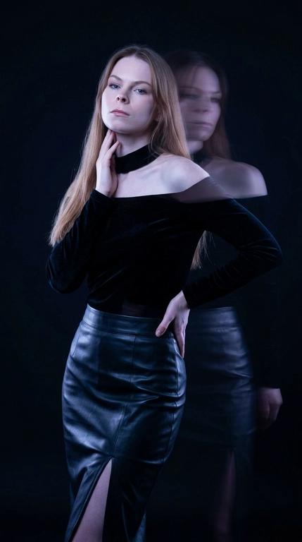 Фотореалистичный fashion-портрет. Женщина стоит вполоборота к камере, корпус плавно изогнут в S-образной линии, одно плечо слегка опущено вперёд, подбородок приподнят. Одна рука поднята к шее и линии ключицы, кончики пальцев мягко касаются области под подбородком, вторая рука согнута в локте и лежит на талии сбоку, локоть отведён в сторону. Голова слегка наклонена назад и в сторону, взгляд направлен на объектив, выражение лица чувственное, уверенное и загадочное.

Тёмная студия практически растворяется в глубоких тенях. Основная фигура остаётся резкой, рядом появляется полупрозрачный смещённый силуэт, создающий эффект двойной экспозиции и призрачного движения. Фон остаётся чистым и тёмным, без световых полос и линий.

Очень низкий ключ освещения, глубокие чёрные тени, минимальное количество света, акцентное холодное освещение по контуру лица, плеч и фигуры, большая часть кадра погружена в полумрак.

Цветокоррекция в стиле мрачной luxury fashion-съёмки: почти чёрный фон, холодные сине-фиолетовые оттенки, высокий контраст, приглушённые света, глубокие бархатные тени, эффект длинной выдержки, motion blur, художественная двойная экспозиция, кинематографичная атмосфера дорогой редакционной съёмки для Vogue, объектив 85mm, f/1.8, высокая детализация, премиальная студийная фотография.


Какая будет прическа?: Не изменять мою прическу

Какая будет одежда?: На женщине приталенный бархатный топ глубокого чёрного оттенка с высоким мягким воротом и открытыми плечами. Внизу длинная кожаная юбка с высокой посадкой и высоким боковым разрезом. Матовый бархат поглощает свет, а кожа даёт холодные отражения от синего неона. Образ выглядит очень дорого, мрачно и чувственно, с эстетикой dark luxury editorial.