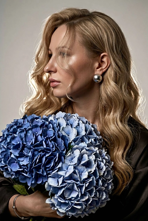Создайте ультрареалистичный портрет, не меняя черты лица, крупным планом 3/4, с мягкой, освещённой кожей, атласной отделкой и естественным блеском, где женщина изображена в профиле. Спокойное и чувственное выражение лица, губы слегка полуоткрыты. Она носит серебряные серьги-шары и простой серебряный браслет на запястье.
Фон нейтральный, в светло-сером тоне, с тёмным, целенаправленным низким угловым освещением, которое подчёркивает контур лица, текстуру кожи и объём волос. Стиль редактирования — высокая мода, чёткий фокус 8K, реалистичная текстура, изысканная и элегантная атмосфера.


Длинные волнистые волосы покрывают часть лица объёмными локонами, выпадающими на лицо и плечо. 

Цветы: Держит в руках огромный букет голубых гортензий