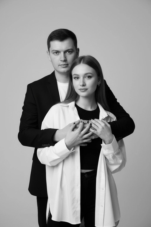 Черно-белый портрет мужчины и женщины в стиле фэшн фотосессии в фотостудии. Не изменяйте лица. Фон нейтральный, однотонный, светло-серый. Атмосфера интимная и спокойная. На переднем плане женщина. Вся композиция выполнена в приглушенной монохромной гамме со студийным освещением, которое подчеркивает текстуру кожи и одежды. Её руки лежат на груди мужчины. Мужчина обнимает женщину сзади, положив свою руку поверх её руки. Она одета в черный топ, белую оверсайз рубашку. Мужчина одет в  классическом черном пиджаке и белой футболке.