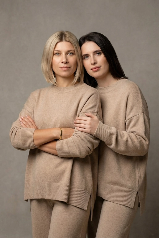 Используй эти два фото мамы и дочери и создай для них студийный портрет в чистом и элегантном стиле. Они стоят рядом, очень близко друг к другу. Дочь мягко обнимает маму за руку и слегка к ней прижимается со спины. Мама сложила руки перед грудью. Образ минималистичный, современный и стильный. На руке у матери тонкий золотой браслет. Профессиональный макияж и прическа.  Не менять черты лица ни на 1 процент. Фон — нейтральный серый с мягкой текстурой. Освещение рассеянное, мягкое, подчёркивающее естественные тона кожи. Атмосфера тёплая, нежная и спокойная, в стиле журнальной съёмки. Обе в свободных стильных бежевых свитерах и брюках.