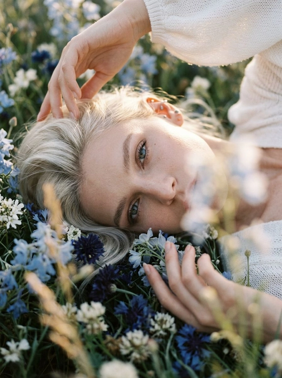 Экстремально крупный, кинематографичный портрет лица человека, лежащего среди густых полевых цветов, камера максимально близко — в кадре преимущественно лицо, черты сохранены без изменений, вокруг море цветов, создающее ощущение мягкого природного кокона, одна рука изящно поднята над головой, вторая мягко касается цветов рядом, на переднем плане сбоку частично размытые, не в фокусе цветы добавляют глубину и лёгкое ощущение движения, одета в летнее белое вязаное платье с объёмными рукавами, мягкая текстура ткани едва читается, естественный мягкий свет подчёркивает кожу и блики в волосах, малая глубина резкости, художественное боке, атмосферный кадр с деликатным плёночным зерном.



Какие будут цветы?: Цветы нежно-голубых, синих и белых оттенков.