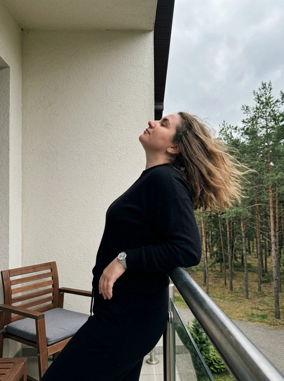 Кэжуал дорогой лайфстайл.

Женщина стоит на балконе отеля, откинула голову назад с закрытыми глазами и слегка опирается на перила. Волосы свободно развеваются.

Фон — балкон загородного отеля в сосновом лесу: светлая штукатурная стена здания, металлические перила с горизонтальными прутьями, деревянное кресло с серой подушкой частично попадает в кадр. За балконом густой сосновый лес и серое облачное небо.

Освещение дневное, мягкое, пасмурное, рассеянное. Фон не размыт и хорошо читается. Вертикальный кадр, камера расположена ниже уровня плеч, съёмка снизу вверх.

Высокая детализация и текстура кожи, shot on iPhone 17 Pro. Эффект плёнки, плёночное зерно, сниженный контраст. Casual smartphone photo, handheld phone shot, quick snapshot, unplanned moment, taken casually without preparation, not meant to be a perfect photo, everyday unpolished look.

Какая будет одежда?: Чёрный спортивный костюм

Доп. характеристики: Женщина 54 размера, черты лица без изменений