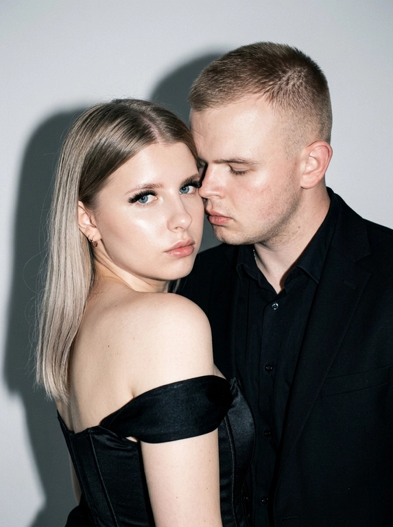 Не меняя черты лица, студийная fashion-съемка пары в минималистичном интерьере с серо-белым фоном, атмосфера гламура и интимной близости, ракурс — крупный портрет по грудь с легким поворотом корпуса, женщина на переднем плане, мужчина позади склоняется к её плечу, поза — она слегка отвернута от камеры с прямым взглядом, он касается лицом её шеи/плеча, одежда — на женщине черный корсет/бандо с открытыми плечами, на мужчине классический черный пиджак и рубашка, макияж — glam (ровный сияющий тон, сильный хайлайтер, четкий контуринг, стрелки, густые ресницы, глянцевые губы), свет — жесткая фронтальная вспышка + мягкий заполняющий свет, выраженные тени позади, эффект flash photography / editorial, холодно-нейтральная цветокоррекция с контрастом и глянцем кожи, высокая детализация, slight film grain, shallow depth of field, съемка на Canon EOS R / 50mm, f/2.0, direct flash, studio look.