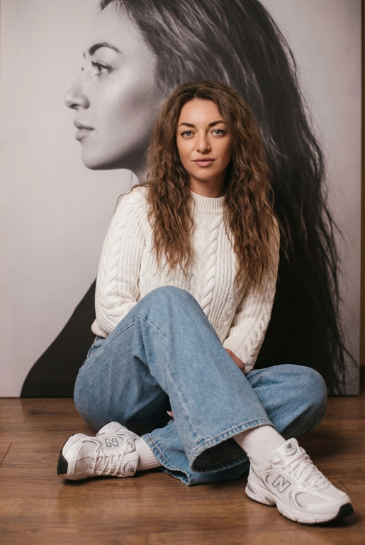   Максимально сохранить внешность, причёску и длину, как на фото.
Женщина должна сидеть на полу в изящной позе, в современном белом джемпере , в широких джинсах и белых носках и белых  кроссовках New Balance.

Её волосы как на фото
Выражение лица, как на фото, она смотрит на камеру .

Фон — тот же её профиль, в мягком крупном чёрно-белом художественном стиле, крупный план сбоку.

Композиция должна быть художественной, мягкой, реалистичной.
Создай изображение, которое выглядит как настоящая фотография.
