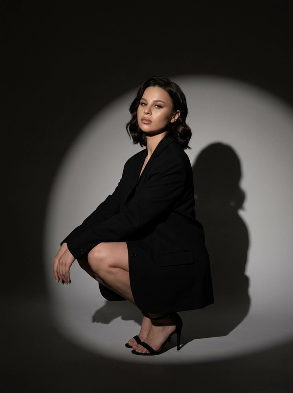 Модная студийная съёмка в стиле fashion editorial. Не менять внешность женщины. Женщина одета в только в чёрный оверсайз пиджак. Профессиональный макияж с акцентом на кожу и глаза. Один направленный прожектор формирует чёткое круглое световое пятно на темно-сером бесшовном гладком фоне и выразительную тень за спиной. Контрастный кинематографичный свет, глубокие тени, драматичный минимализм. Атмосфера — уверенность, сила, эстетика high fashion. высокая детализация кожи, реалистичная текстура, съёмка уровня модного журнала. Средний план. Женщина сидит на корточках, одна рука лежит на колене, корпус слегка наклонён вперёд. Уверенный прямой взгляд в камеру. На ногах чёрные босоножки на тонком каблуке. Чёткая тень позади усиливает драматизм кадра.

Какая будет прическа?: Волосы распущены, уложены в легкие волны.