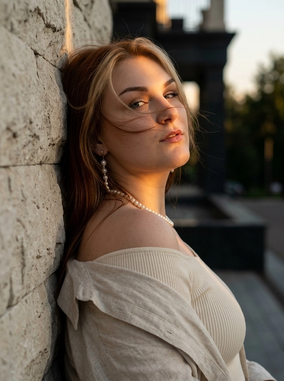 Luxury editorial portrait, кадр по плечи. Женщина стоит вплотную к светлой каменной стене.
Стена находится строго за её спиной и слева в кадре. К стене прижаты верхняя часть спины и левое плечо. Женщина стоит к камере боком, корпус развернут вправо в 3/4, грудь направлена от стены в сторону камеры. Голова резко повернута назад к камере и слегка наклонена вниз. Взгляд в камеру, губы слегка приоткрыты, выражение спокойное, уверенное. На ней молочный плотный топ в рубчик с открытыми плечами, сверху светлая льняная рубашка спущена с плеч. жемчужная нить на шее, длинная серьга с жемчугом. Волосы: несколько прядей слегка раздувает вечерний ветер, перекрывают лицо.
Тёмный закатный golden hour: узкая вертикальная полоса тёплого света освещает один глаз, часть лба, нос, шею и плечо, остальное в тени. Тёмный архитектурный фон мягкий.