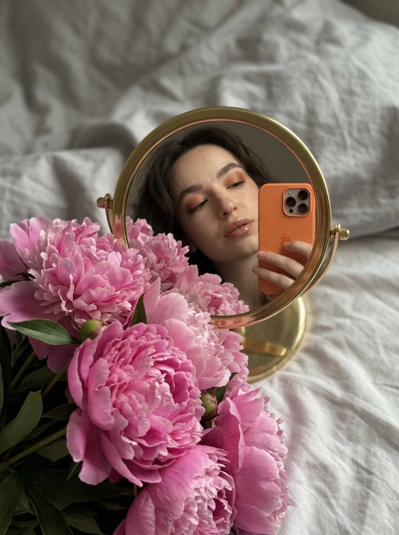 Создай портрет, не меняя черты лица. Фотография в стиле селфи в зеркале, отражающая женщину с безупречным макияжем. На переднем плане — огромный, роскошный букет цветов, которые занимают значительную часть кадра, Женщина держит оранжевый iPhone 17 PRO. Зеркало компактное круглой формы в золотой оправе лежит на белой кровати, женщина склоняется над ним, ракурс снизу. 
Атмосфера и детали:
Фон — мягкое серое постельное белье с естественными складками, создающее уютную домашнюю обстановку. Освещение мягкое, приглушенное, с акцентом на насыщенный цвет цветов. Композиция кадра динамичная: зеркало расположено под углом, создавая интересную перспективу и глубину.
Технические параметры:
Высокая детализация, реализм, текстура лепестков цветов и ткани, глубина резкости (bokeh) на заднем плане, эстетика Pinterest, 8k, кинематографичное качество.

Букет: Роскошные розовые пионы