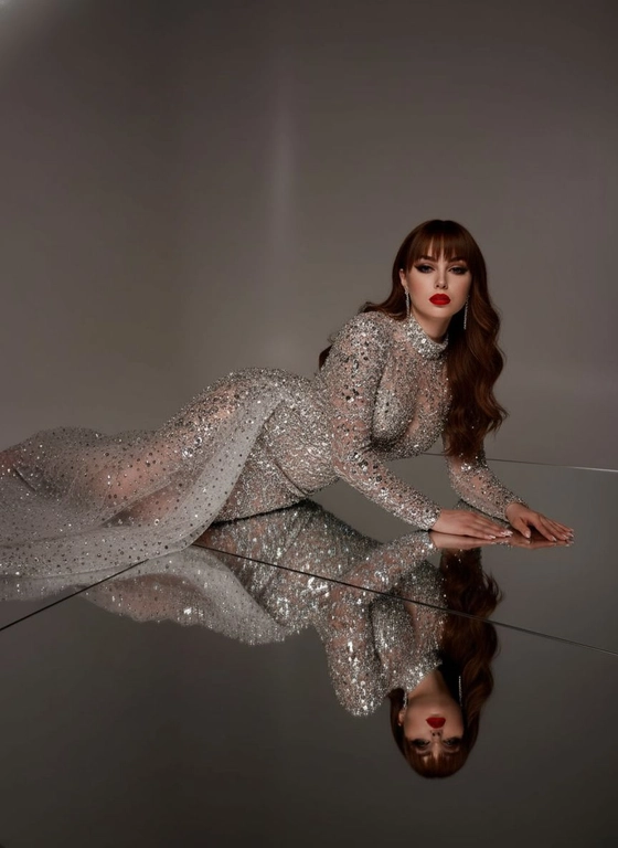 Это изысканный портрет женщины, лежащей боком на зеркальной поверхности, создавая идеальное отражение всего ее тела. На ней элегантное, облегающее платье от кутюр, полностью расшитое серебряными стразами, кристаллами и сверкающими деталями, которые роскошно ловят свет. Платье имеет длинные рукава, высокий ворот и струящийся шлейф из прозрачного тюля, усыпанного стразами на юбке, источающий гламур и изысканность. У нее длинные, волнистые волосы, которые нежно падают на ее плечи. Ее макияж безупречен с яркими темными smoky eyes и черной подводкой для глаз, длинными, объемными ресницами, безупречной кожей и пухлыми губами с изысканной, яркой красной помадой. На ней сдержанные, но элегантные серьги, дополняющие гламурный образ. Сцена драматически освещена темной виньеткой, подчеркивающей блеск платья и контраст между ее телом и отражением в зеркале. Минималистичный фон в нейтральных тонах ещё больше подчёркивает харизму модели и богатство деталей наряда. Общая атмосфера создаёт ощущение роскоши, элегантности и изысканности, отражая моду и высокую моду. Сохранены те же черты лица без каких-либо изменений, тот же тон кожи и цвет волос. Качественный маникюр, профессиональное студийное качество изображения.