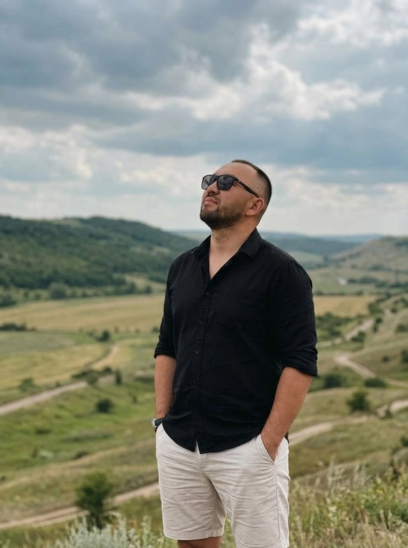 Создай фото, не меняя черты лица. Мужчина стоит на фоне холмистого природного пейзажа под облачным небом. Снимок выполнен в стиле спокойной lifestyle-фотографии с естественной композицией и мягким дневным светом. Атмосфера умиротворённая, свободная и немного кинематографичная. Цветовая гамма приглушённая, с землистыми оттенками, натуральной зеленью и мягким контрастом.

На мужчине чёрная рубашка с коротким рукавом, застёгнутая на несколько пуговиц, и светлые шорты. Руки расслабленно находятся в карманах. Он слегка запрокидывает голову и смотрит вверх. На лице тёмные солнцезащитные очки. Фон слегка размыт, акцент на человеке, реалистичная кожа, естественная поза, высокая детализация, без глянцевого AI-эффекта.