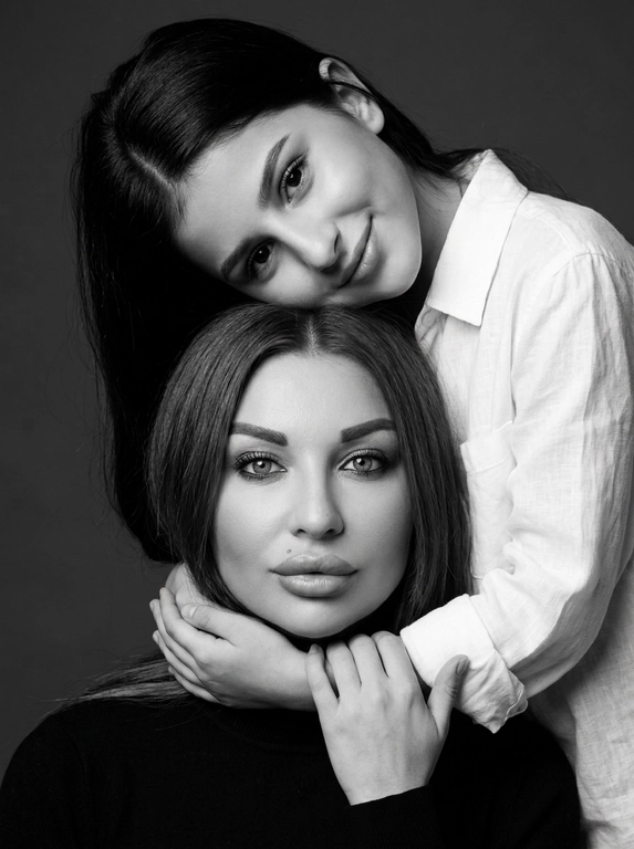 Родитель и ребёнок с внешностью как на приложенных фото, художественная семейная студийная фотосессия. Композиция — вертикальный портрет, крупный план лиц. Родитель внизу смотрит прямо в камеру, лицо фронтально. Ребёнок сверху, голова повернута набок, щека лежит на голове родителя, смотрит в камеру с нежной улыбкой. Рука ребёнка обнимает родителя за шею, видна в кадре, родитель держит руку ребёнка своей рукой.
Одежда: ребёнок в белой льняной рубашке, родитель в чёрной водолазке. 
Фон — тёмно-серый почти чёрный однотонный студийный, создаёт контраст. Освещение — мягкое боковое студийное, создаёт объём и глубину.
Чёрно-белая художественная фотография с выразительными контрастами. Атмосфера нежная, интимная, спокойная.