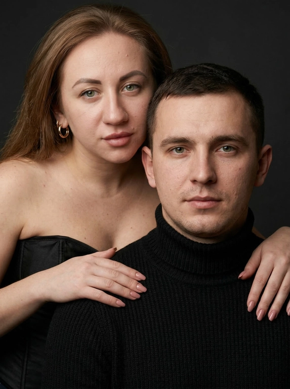 Гиперреалистичный студийный fashion-портрет молодой пары по загруженному референсу, 100% сходство лиц. Черты лица не изменять. Крупный план, кадр по плечи. Мужчина, спокойный уверенный взгляд прямо в камеру, естественное выражение лица, одет в черный трикотажный свитер с высоким воротом. Женщина стоит позади мужчины и нежно обнимает его, ее руки лежат на его плечах. Ее лицо расположено рядом с его лицом, она повернута слегка боком, голова немного наклонена вперед, взгляд направлен прямо в камеру. У женщины выразительные глаза с длинными густыми черными ресницами, естественный clean-glam макияж, натуральные брови, теплый оливковый оттенок кожи, небольшие серьги-кольца. Композиция плотная и интимная, их лица расположены очень близко друг к другу, современный модный портрет пары. Женщина одета в черный корсет. Кожа максимально натуральная: живая текстура кожи, видимые микропоры, subtle skin glow, естественный мягкий блеск кожи, natural skin microtexture, skin translucency, micro skin details, легкие естественные несовершенства кожи, без ретуши и без эффекта пластика. Освещение: мягкий студийный beauty lighting, рассеянный ключевой свет спереди и немного сбоку, мягкие глубокие тени для объема лица, деликатные световые блики на коже. Фон: глубокий темный студийный фон, темно-графитовый или почти черный, создающий контраст с кожей и лицами. Стиль: luxury fashion editorial photography, high fashion portrait, ultra photorealistic, 85mm portrait lens, shallow depth of field, creamy bokeh, cinematic soft lighting, RAW photo, extremely detailed, fine hair strands, natural skin pores, professional photography, photorealism, 8k, high dynamic range.