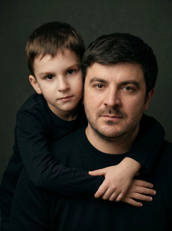 Создай портретный студийный кадр из фотосессии отца и сына. Сын обнимает отца сзади, лежа подбородком на его плече, руки мягко сомкнуты на груди мужчины. Оба смотрят прямо в камеру. Естественные спокойные эмоции, лёгкая серьёзность во взгляде.
Одежда — однотонные чёрные лонгсливы или свитера без принтов. Тёмный нейтральный фон (глубокий серо-оливковый или графитовый). Мягкий направленный студийный свет с лёгкой теневой глубиной, акцент на лицах, тёплая естественная цветокоррекция, высокая детализация кожи, натуральная текстура без излишней ретуши.
Кадр — крупный или средний план, плотная композиция, лица расположены близко друг к другу. Атмосфера — тёплая, камерная, эмоциональная, ощущение близости и доверия. Профессиональная съёмка, 85mm lens, shallow depth of field, cinematic portrait, ultra realistic.