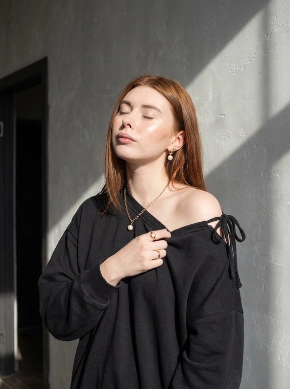 Используй загруженное фото для лица, строго не меняя черты лица, пропорции, возраст, текстуру кожи и волосы. Минималистичный портрет в интерьере с естественным светом, женщина стоит у серой текстурированной оштукатуренной стены, голова слегка запрокинута назад, глаза закрыты, лицо повернуто к источнику света, правая рука держит ткань толстовки у груди, левая рука расслабленно опущена вдоль тела. Оверсайз толстовка с капюшоном спущена с правого плеча, видны завязки, на шее тонкая золотая цепочка с кулоном с жемчужиной, золотые серьги среднего размера с жемчугом, золотое кольцо на пальце. Естественный макияж с деликатным скульптурированием, влажное свечение кожи, кожа ультрареалистичная без эффекта пластика и чрезмерной ретуши. Жёсткий направленный солнечный свет из окна создаёт резкие графичные тени на стене и лице, контрастное освещение, слева тёмный дверной проём добавляет глубину пространству. Съёмка на 50mm f/2.0, малая глубина резкости, естественный оконный свет, выраженные тени, цветовая гамма бежевый, золото, серый, баланс тёплых и холодных оттенков, minimalist editorial, фотореализм высокого качества.

Какой цвет толстовки?: Цвет толстовки чёрный.