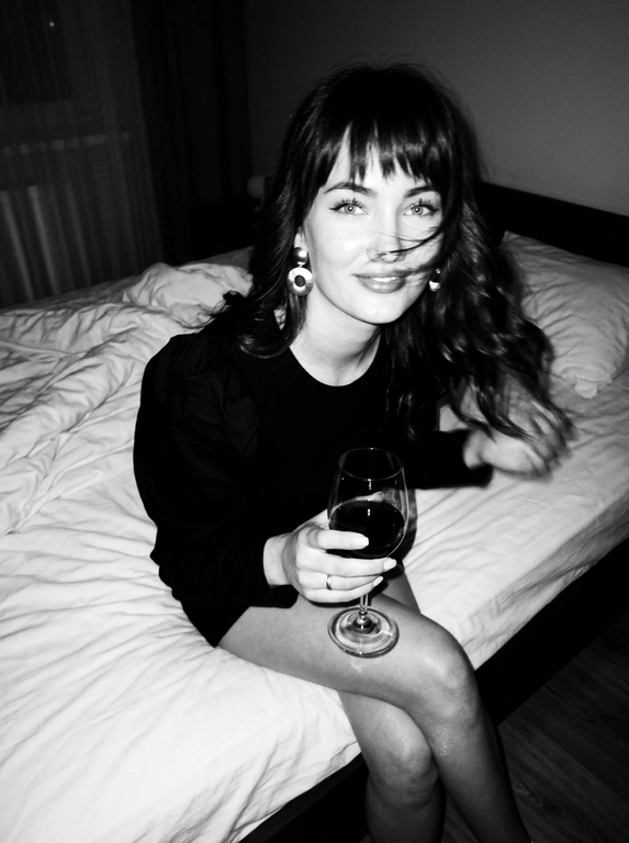 Create a portrait, transfer the face from the reference photo without changing facial features, proportions, or expression, preserve full identity. Aesthetic black-and-white nighttime Y2K photograph captured with direct on-camera flash, shot in motion.
A woman is sitting on the edge of a white, slightly rumpled bed, the frame taken from a slightly elevated angle. She wears a stylish black top with voluminous sleeves and large round statement earrings. In one hand she holds an elegant glass of dark wine. Her body is subtly moving, creating natural motion blur across the face and shoulders, as if the moment was caught mid-gesture. The gaze is languid and directed toward the camera, with a soft, intimate smile. Hair falls in large waves with loose strands drifting across the face, slightly displaced by movement. Lighting is harsh flash against deep night shadows, high contrast with blown highlights and strong falloff, visible film grain. Skin texture remains real and alive, no smoothing, enhanced by flash glare. The mood is high-fashion editorial, Parisian chic, old-money sensuality, nighttime melancholy, spontaneous and cinematic, shot in a raw 35mm film aesthetic with pronounced motion blur and lived-in realism.