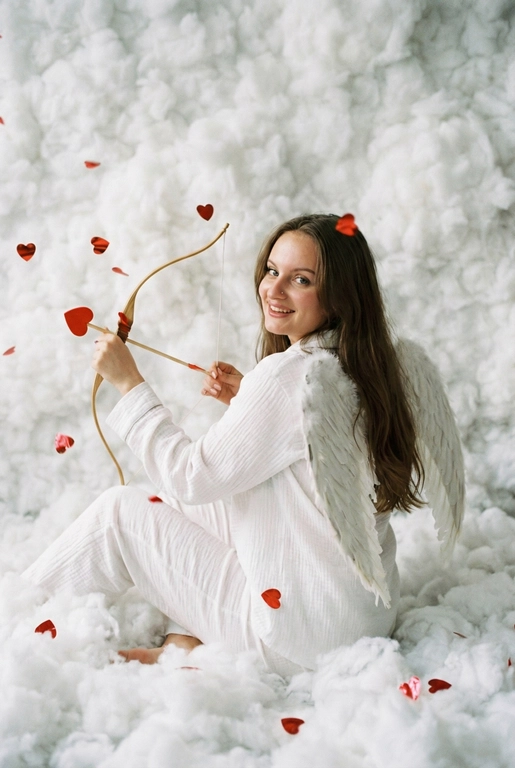 Использовать реальные черты лица человека с референса, естественную текстуру кожи и настоящий цвет и длину волос. Личность должна оставаться неизменной.

Y2K. Сказочная сцена с купидоном, сфотографированная как реальный момент.
Человек сидит среди плотных, пушистых белых облаков, которые полностью заполняют передний, средний и задний план, образуя бесшовную белую среду с многослойной глубиной.

Поза вполоборота: корпус слегка повернут вбок, одно плечо выдвинуто вперед, как будто человек натягивает и отпускает небольшой лук с красной стрелой в форме сердца; руки подняты с легким напряжением, указывающим на движение при выстреле.

Несмотря на поворот тела, голова повернута к объективу, взгляд направлен прямо в камеру. Выражение лица радостное и игривое, с мягкой улыбкой.

Наряд — простая белая пижама: свободная рубашка и брюки из муслина.
На спине — крылья из белых перьев, настоящие и фактурные, с четкими бликами по краям каждого пера.

Вокруг объекта в воздухе хаотично летают только конфетти в форме сердечек — маленькие красные сердечки из глянцевой фольги, расположенные на разном расстоянии: часть в переднем плане, часть растворяется среди облаков.

Без рассеянного света, без мягкого свечения, без студийного глоу.
Текстура кожи реалистичная и детализированная, без сглаживания и без фильтров.
Тонкая зернистость, волшебное, но искреннее ощущение Y2K-редакции — выглядит как фотография, а не визуализация.