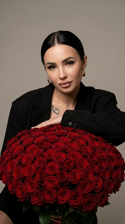 Ультрареалистичный fashion-портрет в эстетике luxury editorial. Крупный план по пояс. Женщина слегка наклоняется над огромным роскошным букетом, который полностью заполняет передний план кадра. Она смотрит прямо в камеру с мягким, уверенным взглядом.
На женщине чёрный оверсайз-пиджак премиального кроя с чёткой линией плеч. 
Макияж luxury soft glam: длинные густые ресницы с лисьим эффектом, чёрные графичные стрелки, ламинированные брови, лёгкий контуринг, персиковые румяна, золотистый хайлайтер, губы с чётким нюдово-коричневым контуром и глянцевым блеском.
Фон минималистичный, нейтральный, тёплых бежево-серых оттенков, без лишних деталей. Освещение — мягкий студийный свет в сочетании с прямой вспышкой камеры, создающий выразительный контраст, естественные блики на коже, лепестках роз и ткани пиджака. Глубокие, но мягкие тени подчёркивают объём и атмосферу дорогой журнальной съёмки.
Максимальная детализация: текстура кожи с видимыми порами, отдельные пряди волос, бархатистые лепестки, фактура ткани пиджака. Живой RAW-эффект, лёгкое плёночное зерно, без ретуши, без пластиковой кожи, без AI-эффекта.
Ultra realistic RAW photo, high fashion editorial, luxury portrait, oversized bouquet of deep red roses, black oversized blazer, direct flash photography, cinematic contrast, authentic skin texture, visible pores, realistic hair strands, premium magazine aesthetic, 8K, sharp focus, no beauty filter, no plastic skin, no AI artifacts.

Какие будут цветы?: Букет из сотен тёмно-красных роз

Какая будет прическа?: Волосы собраны в аккуратный низкий гладкий пучок с ровным центральным пробором. Корни идеально приглажены, без выбивающихся прядей, создавая чистый и строгий luxury-силуэт.