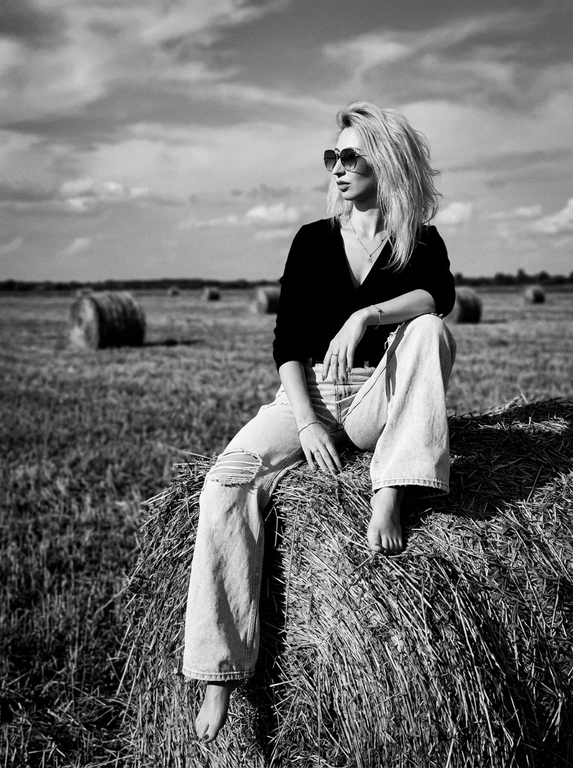 Чёрно-белая high-fashion editorial фотография в сельской местности, женщина с внешностью как на референсе, черты лица не изменять, композиция — более общий план (wide shot), камера на среднем расстоянии, фигура полностью в кадре с окружающим пространством, она сидит на большом тюке сена в поле, корпус слегка развернут, одна нога свободно опущена вниз, вторая согнута, создавая расслабленную, но выразительную позу, руки естественно лежат на колене и на поверхности тюка, взгляд направлен в сторону, профиль, выражение спокойное и уверенное, одежда: чёрный топ с V-образным вырезом и объёмные светлые широкие джинсы с потёртостями и рваными элементами, аксессуары: крупные солнцезащитные очки, минималистичные украшения, фон: открытое поле с несколькими тюками сена на разной глубине, создающими перспективу, линия горизонта ниже центра кадра, чтобы подчеркнуть небо, освещение: жёсткий естественный дневной свет с мягкими облаками, формирует выразительные тени и контраст, стиль: кинематографический, плёночное зерно, высокая детализация, реалистичная текстура кожи и ткани, редакционный fashion стиль, акцент на атмосфере и пространстве кадра


Какая будет прическа?: НЕ изменять мою прическу