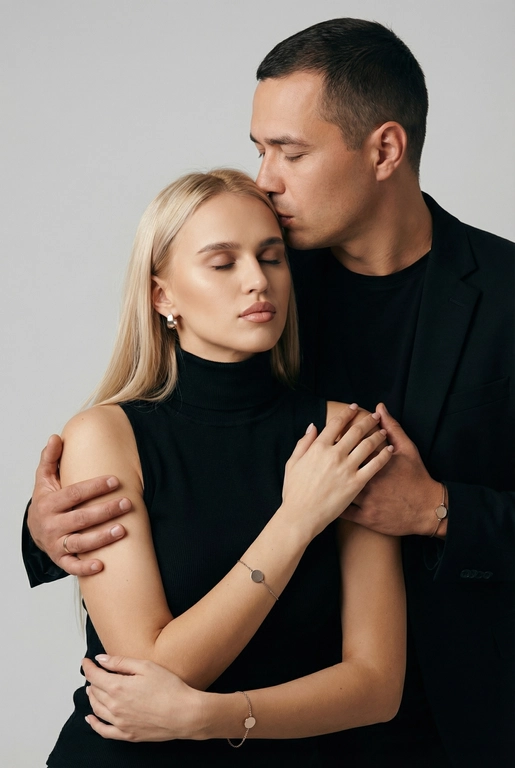   Мужчина и женщина позируют в студии в объятиях. Мужчина стоит позади женщины, обнимая её обеими руками; его тело повернуто к ней, голова слегка наклонена вниз, губы касаются её лба. Женщина немного отклоняет голову назад к его плечу, её глаза закрыты. Кадр — средний, от талии до головы, снят на уровне глаз. Композиция плотная, акцентирует линии рук и соприкосновение лиц. Женщина стоит, прямо обращённая к камере.
На ней облегающий чёрный топ без рукавов с высоким воротом, из плотной матовой ткани.
Мужчина одет в чёрную футболку и чёрную неброскую куртку. На обоих запястьях — тонкие металлические браслеты с минималистичными круглыми подвесками, а женщина носит небольшие светлые металлические серьги с гладкой поверхностью.
Обстановка минималистичная: однотонный светло-серый фон без текстуры.
Освещение — мягкий студийный свет: рассеянный фронтальный и боковой свет создаёт плавные переходы света и тени на лицах и руках. Тени мягкие, средней глубины. Контраст умеренный, без жёстких бликов. Свет равномерно распределён по фигурам, фон подсвечен мягко, без выраженных градиентов.
Съёмка на портретный объектив 85 мм, малая глубина резкости с чётким фокусом на лицах и руках; фон слегка размыт, текстуры кожи и ткани высоко детализированы. Это минималистичный студийный портрет, высокодетализированный и фотореалистичный.
Минималистичное романтическое студийное настроение, нейтральные тёплые тона кожи, низкая–средняя насыщенность, мягкое рассеянное освещение, средний контраст с нежными тенями, плавные переходы бликов, естественная передача кожи, чистый светлый фон, тонкий тональный баланс, современная мягкая редакционная цветокоррекция. Не изменять мою прическу