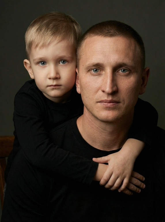 Создай портретный студийный кадр из фотосессии отца и сына. Сын обнимает отца сзади, лежа подбородком на его плече, руки мягко сомкнуты на груди мужчины. Оба смотрят прямо в камеру. Естественные спокойные эмоции, лёгкая серьёзность во взгляде.
Одежда — однотонные чёрные лонгсливы или свитера без принтов. Тёмный нейтральный фон (глубокий серо-оливковый или графитовый). Мягкий направленный студийный свет с лёгкой теневой глубиной, акцент на лицах, тёплая естественная цветокоррекция, высокая детализация кожи, натуральная текстура без излишней ретуши.
Кадр — крупный или средний план, плотная композиция, лица расположены близко друг к другу. Атмосфера — тёплая, камерная, эмоциональная, ощущение близости и доверия. Профессиональная съёмка, 85mm lens, shallow depth of field, cinematic portrait, ultra realistic.

