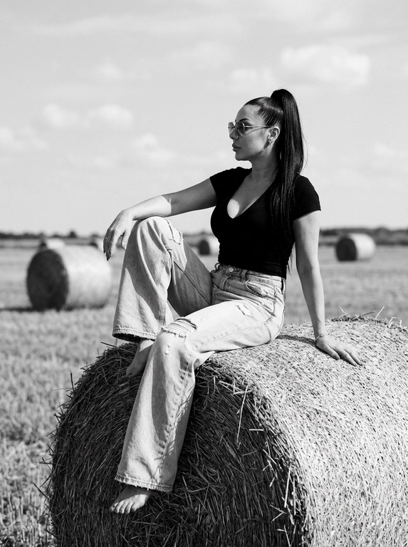 Чёрно-белая high-fashion editorial фотография в сельской местности, женщина с внешностью как на референсе, черты лица не изменять, композиция — более общий план (wide shot), камера на среднем расстоянии, фигура полностью в кадре с окружающим пространством, она сидит на большом тюке сена в поле, корпус слегка развернут, одна нога свободно опущена вниз, вторая согнута, создавая расслабленную, но выразительную позу, руки естественно лежат на колене и на поверхности тюка, взгляд направлен в сторону, профиль, выражение спокойное и уверенное, одежда: чёрный топ с V-образным вырезом и объёмные светлые широкие джинсы с потёртостями и рваными элементами, аксессуары: крупные солнцезащитные очки, минималистичные украшения, фон: открытое поле с несколькими тюками сена на разной глубине, создающими перспективу, линия горизонта ниже центра кадра, чтобы подчеркнуть небо, освещение: жёсткий естественный дневной свет с мягкими облаками, формирует выразительные тени и контраст, стиль: кинематографический, плёночное зерно, высокая детализация, реалистичная текстура кожи и ткани, редакционный fashion стиль, акцент на атмосфере и пространстве кадра


Какая будет прическа?: Гладкие волосы собраны в высокий хвост
