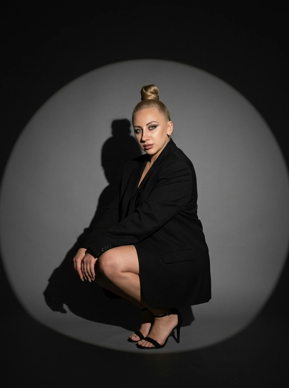 Модная студийная съёмка в стиле fashion editorial. Не менять внешность женщины. Женщина одета в только в чёрный оверсайз пиджак. Профессиональный макияж с акцентом на кожу и глаза. Один направленный прожектор формирует чёткое круглое световое пятно на темно-сером бесшовном гладком фоне и выразительную тень за спиной. Контрастный кинематографичный свет, глубокие тени, драматичный минимализм. Атмосфера — уверенность, сила, эстетика high fashion. высокая детализация кожи, реалистичная текстура, съёмка уровня модного журнала. Средний план. Женщина сидит на корточках, одна рука лежит на колене, корпус слегка наклонён вперёд. Уверенный прямой взгляд в камеру. На ногах чёрные босоножки на тонком каблуке. Чёткая тень позади усиливает драматизм кадра.

Какая будет прическа?: Не изменять мою прическу