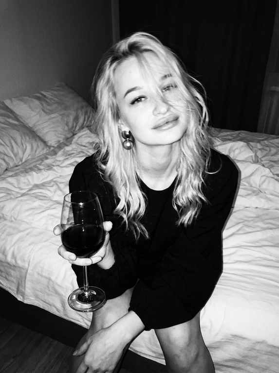 Create a portrait, transfer the face from the reference photo without changing facial features, proportions, or expression, preserve full identity. Aesthetic black-and-white nighttime Y2K photograph captured with direct on-camera flash, shot in motion.
A woman is sitting on the edge of a white, slightly rumpled bed, the frame taken from a slightly elevated angle. She wears a stylish black top with voluminous sleeves and large round statement earrings. In one hand she holds an elegant glass of dark wine. Her body is subtly moving, creating natural motion blur across the face and shoulders, as if the moment was caught mid-gesture. The gaze is languid and directed toward the camera, with a soft, intimate smile. Hair falls in large waves with loose strands drifting across the face, slightly displaced by movement. Lighting is harsh flash against deep night shadows, high contrast with blown highlights and strong falloff, visible film grain. Skin texture remains real and alive, no smoothing, enhanced by flash glare. The mood is high-fashion editorial, Parisian chic, old-money sensuality, nighttime melancholy, spontaneous and cinematic, shot in a raw 35mm film aesthetic with pronounced motion blur and lived-in realism.