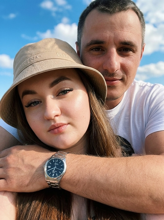 Кинематографичный кадр крупным планом (selfie style), пара в объятиях (классическое "обнимашки сзади") на фоне ярко-голубого неба с облаками.  Женщина на ней бежевая панама с брендовой надписью. Её кожа выглядит сияющей, подсвеченной мягким естественным солнечным светом. Выражение лица спокойное, глубокий взгляд, направленный прямо в камеру. Мужчина стоит строго позади девушки и обнимает её сзади, одна его рука обнимает её за плечи/шею спереди — предплечье лежит поперёк её ключиц, кисть свободно свисает с её плеча, на запястье классические серебряные часы с черным циферблатом. Мужчина в белой футболке, выражение лица доброжелательное, его взгляд прямо в объектив. Ракурс сверху, камера сфокусирована на лице девушки. Высокий контраст, насыщенные цвета, мягкая прорисовка деталей кожи. Эффект пленочной фотографии, легкое зерно, атмосфера летнего дня, эстетика социальных сетей. Телефона не видно в кадре.
