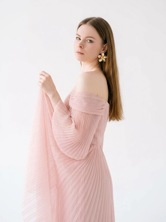 Студийный портрет женщины, выполненный в стиле fashion-съемки. Фон белый, без каких-либо отвлекающих деталей. Свет мягкий, рассеянный, создающий ощущение свежести и легкости. Цветовая гамма нейтральная, с преобладанием пастельных тонов.
На женщине надето платье из полупрозрачной плиссированной ткани . Платье открывает одно плечо. На ухе видна массивная золотая серьга в форме цветка.
Женщина стоит полубоком к камере, повернув голову назад через плечо. Взгляд направлен в камеру. Поза расслабленная и уверенная. Одна рука приподнята и частично придерживает ткань платья на плече.

Цвет платья: Платье светлого розового оттенка