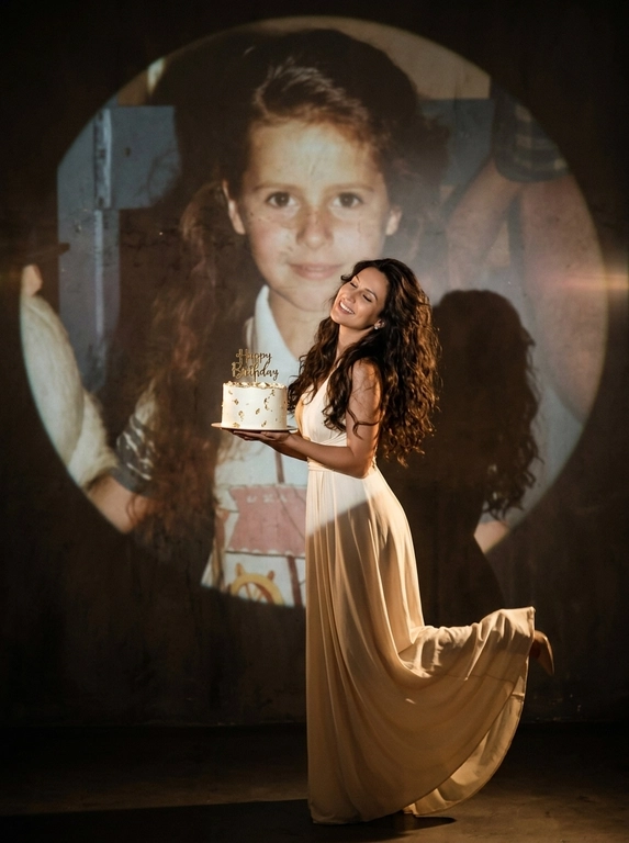 Create ultra-realistic cinematic birthday photoshoot, elegant studio with a dark background and a large circular spotlight projection on the wall. Inside the spotlight, a faded vintage childhood photo of the same girl(the reference image 2nd given)appears as a nostalgic memory, slightly transparent with old photo texture and soft blur. In the foreground, the present-day version of the same model (the reference image 1st given)  holding a white birthday cake decorated with gold accents with a “Happy Birthday". 

If the model is a woman, then she is wearing a cream long gown. If the model is a man, he is wearing a suit.

her head tilted back, eyes closed, natural smile,  cinematic shadow cast on the wall, warm soft spotlight lighting.

If the model was a woman, she additionally lifted her leg in an elegant pose.